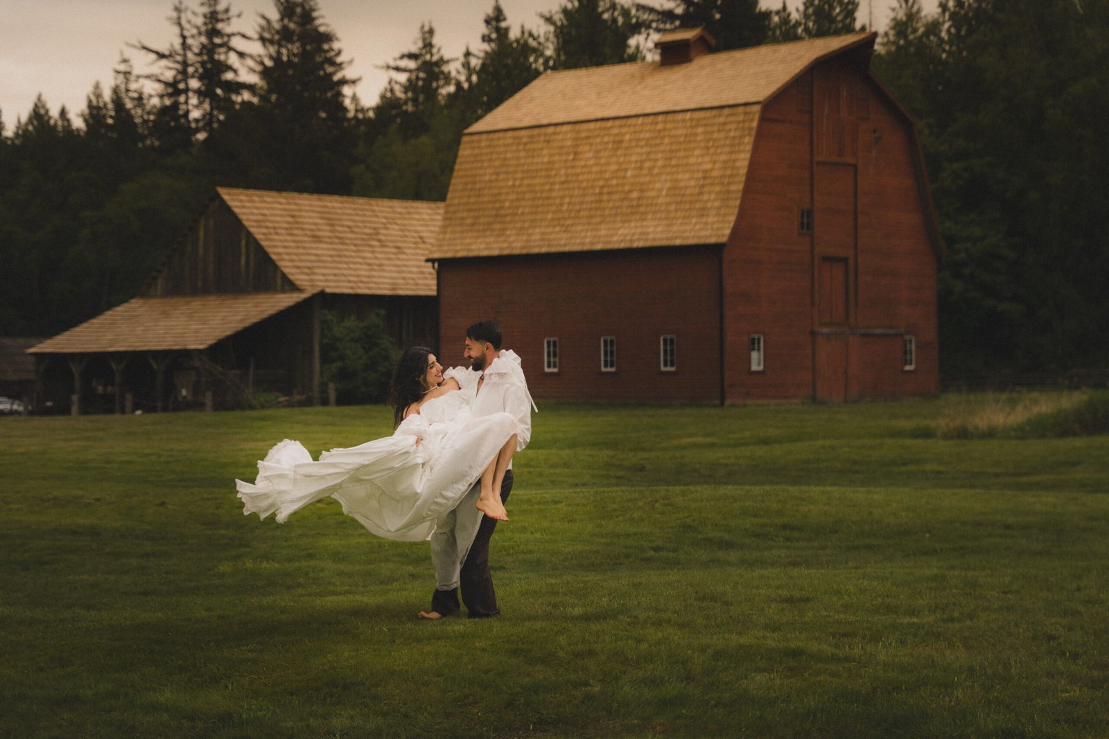 Groom lifting the bride in front of a red barn on a grassy field
