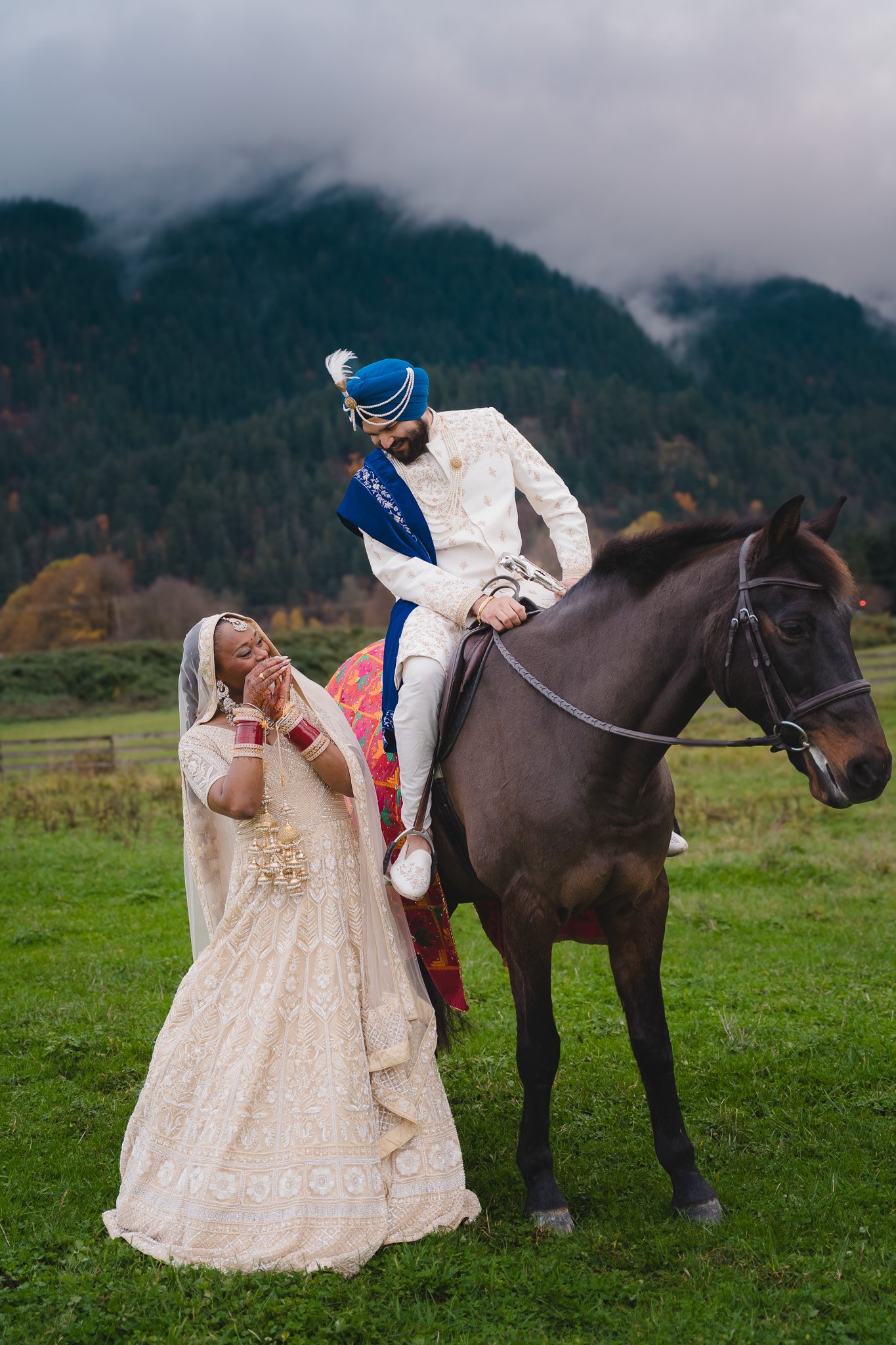 Groom in a blue turban on horseback beside his bride in an embroidered lehenga, mountains behind